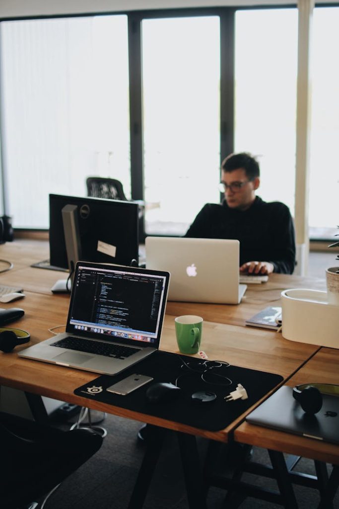 man-in-black-shirt-sits-behind-desk-with-computers-2102416 A web developer working on code in a modern office setting with multiple devices.